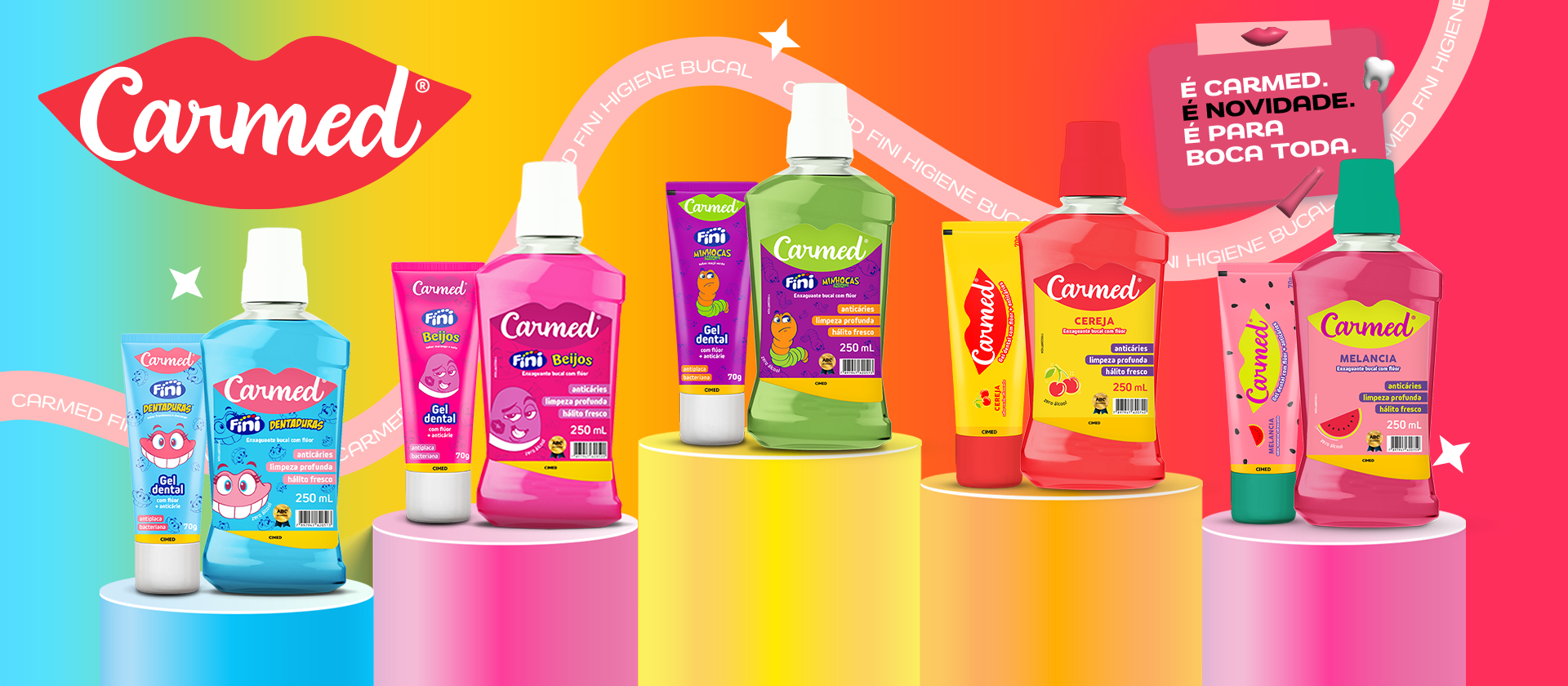 Banner colorido de lançamento da linha Carmed Higiene Bucal. O fundo traz um degradê vibrante em tons de azul, amarelo, laranja e rosa, atravessado por uma fita com o texto 'Carmed Fini Higiene Bucal'. No canto superior esquerdo está o logo da Carmed em formato de boca. No canto superior direito, um post-it rosa diz: 'É Carmed. É novidade. É para boca toda.'. Ao centro, apoiados sobre pedestais cilíndricos coloridos, estão cinco pares de produtos contendo gel dental e enxaguante bucal nos sabores: Fini Dentaduras (azul), Fini Beijos (rosa), Fini Minhocas (verde), Cereja (vermelho) e Melancia (rosa e verde).